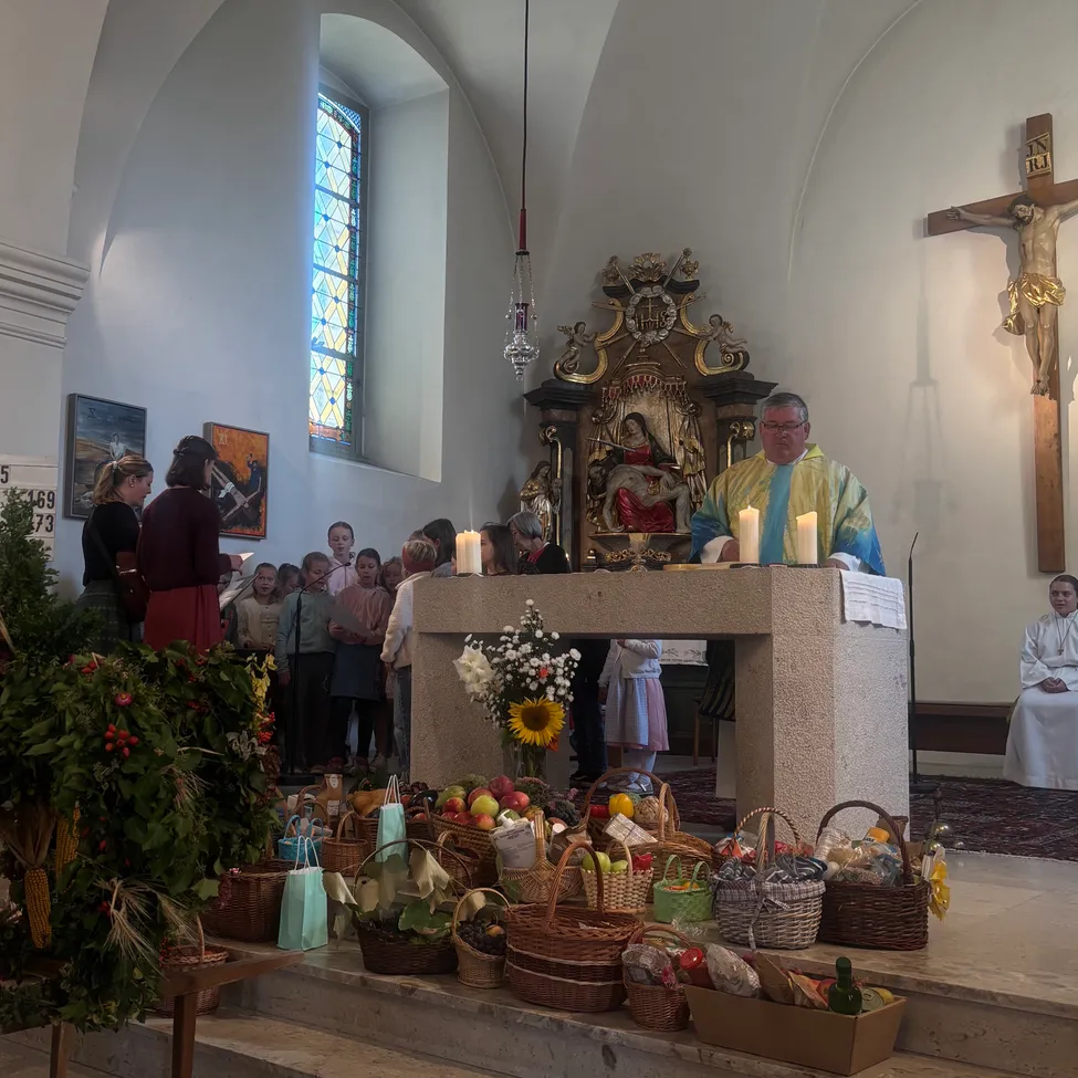 Ein Priester steht am Altar in einer Kirche, umgeben von Körben mit Früchten und Blumen, mit einem Kreuz an der Wand hinter ihm.