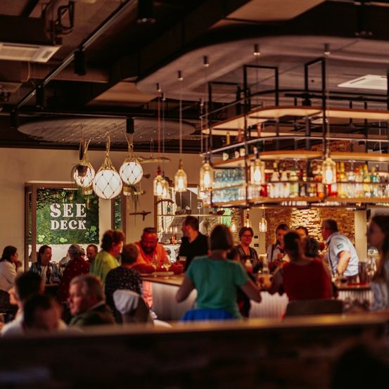 Eine lebhafte Bar mit einer Menschenmenge, die sich um die Theke und Barhocker versammelt. Dekorative Lampen hängen von der Decke.