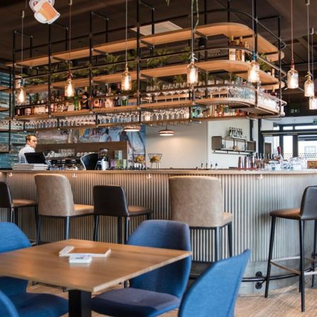 Das Innere einer modernen Bar mit einer langen Theke, mehreren Stühlen und Tischen. Ein Barkeeper steht hinter der Theke, und Regale mit Flaschen und Gläsern hängen von der Decke.