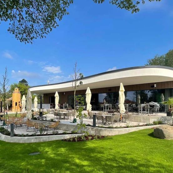 Ein Outdoor-Essbereich mit Tischen und Stühlen, umgeben von Grün und Bäumen. Das Gebäude hat große Fenster und ein modernes Design.