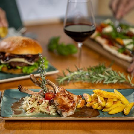 Auf einem Holztisch befinden sich ein Teller mit Schweinekoteletts, Sauerkraut und Pommes, ein Glas Wein und ein Burger mit grünen Beilagen.