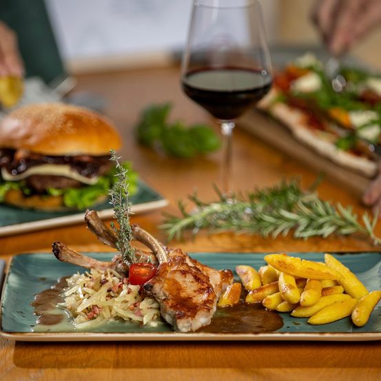 Auf einem Holztisch befinden sich ein Teller mit Schweinekoteletts, Sauerkraut und Pommes, ein Glas Wein und ein Burger mit grünen Beilagen.