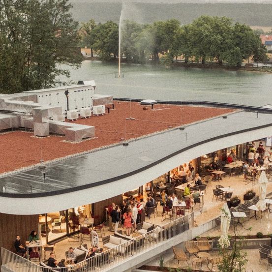 Eine Luftaufnahme eines modernen Dachcafés mit vielen Gästen, die an Tischen sitzen. Das Café hat ein rotes Ziegeldach und Solarmodule. Bäume und ein Fluss sind im Hintergrund zu sehen.