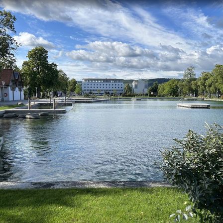 Eine ruhige Seeszene mit einem großen Gebäude im Hintergrund, umgeben von Bäumen und Rasenflächen, unter einem blauen Himmel mit verstreuten Wolken.