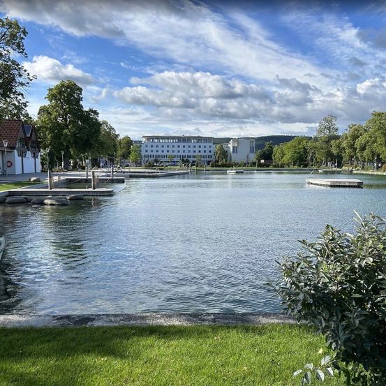Eine ruhige Seeszene mit einem großen Gebäude im Hintergrund, umgeben von Bäumen und Rasenflächen, unter einem blauen Himmel mit verstreuten Wolken.