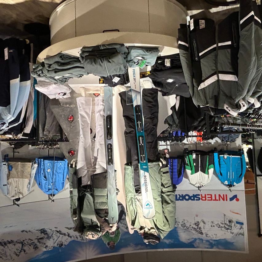 Ein Skiladen zeigt Skijacken und -hosen an Haken hängend, zusammen mit Skiern. Einige Artikel haben Preisschilder. Der Laden hat ein großes Schild mit Bergen und Schnee.