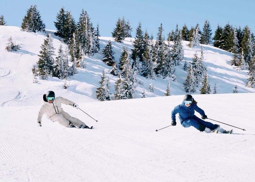Zwei Skifahrer rutschen einen schneebedeckten Berg hinunter, an dem Kiefern im Schnee liegen, beide tragen Helme und Skianzüge.