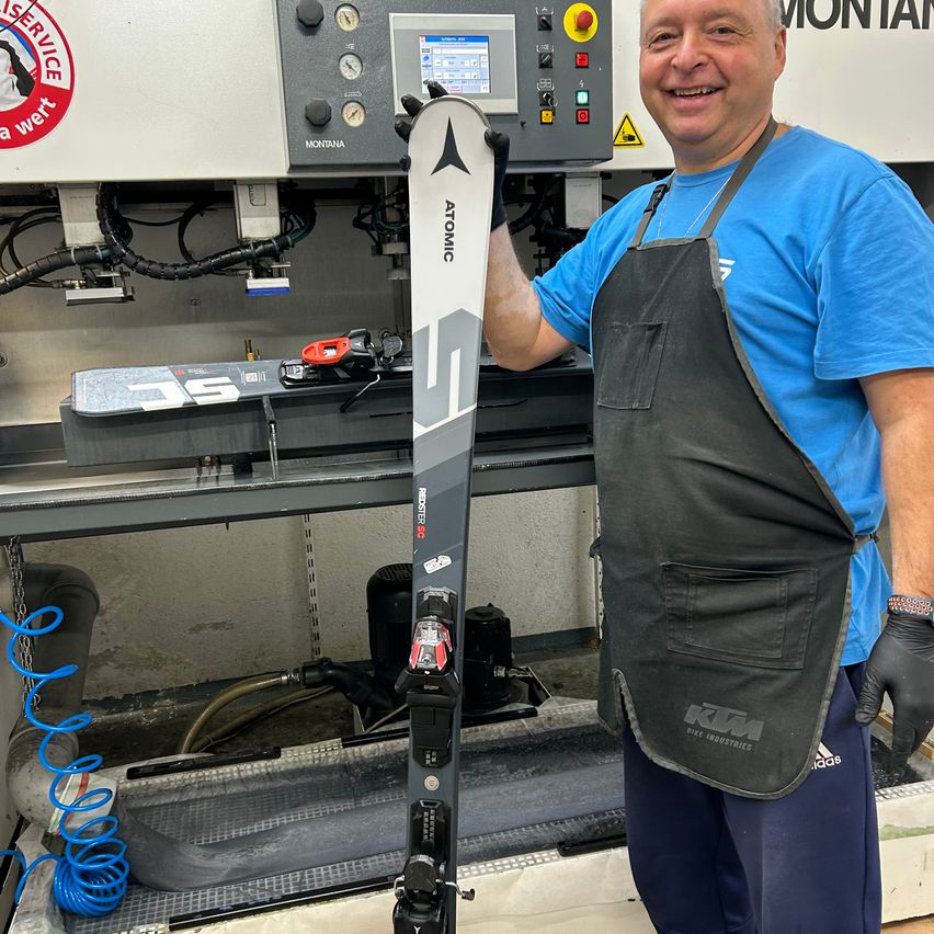 Ein Mann in einem blauen Hemd und einer Schürze steht in einer Werkstatt, lächelt und hält ein Paar Atomic-Skier in der Hand. Hinter ihm ist eine große Maschine mit Bildschirm und Bedienfeld zu sehen.