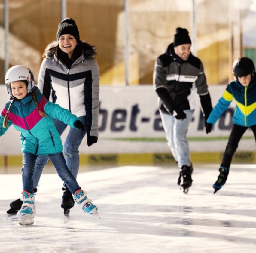 Eine Familie ist auf einer Eisbahn beim Eislaufen. Mutter, Vater und zwei Kinder lächeln alle. Die Eisbahn hat eine weiße Barriere mit Text.