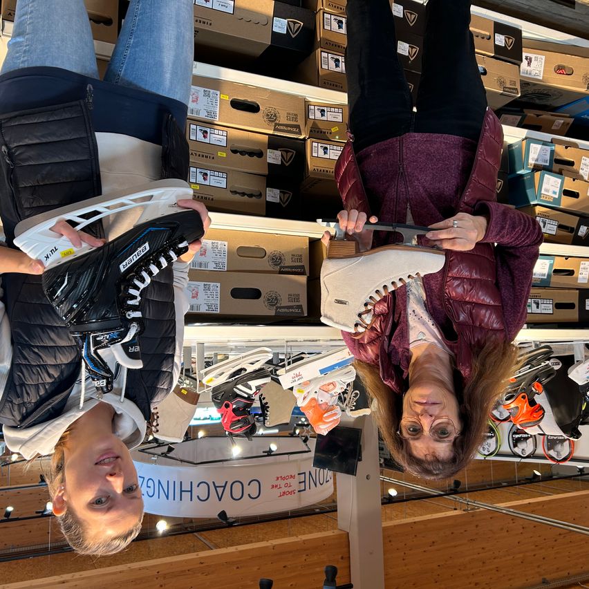 Zwei Frauen sind kopfüber in einem Schuhgeschäft. Eine hält einen Schlittschuh, die andere einen Schuh. Sie lächeln und scheinen für ein Foto zu posieren.