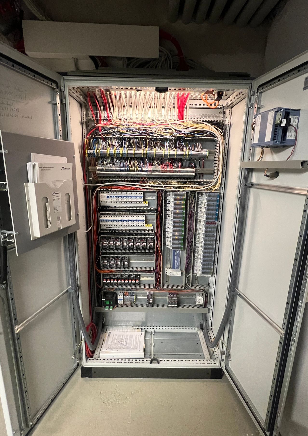 Ein geöffnetes elektrisches Schaltschrank mit zahlreichen Kabeln und Schaltkreisen. Papiere und Dokumente sind an der Seite befestigt.
