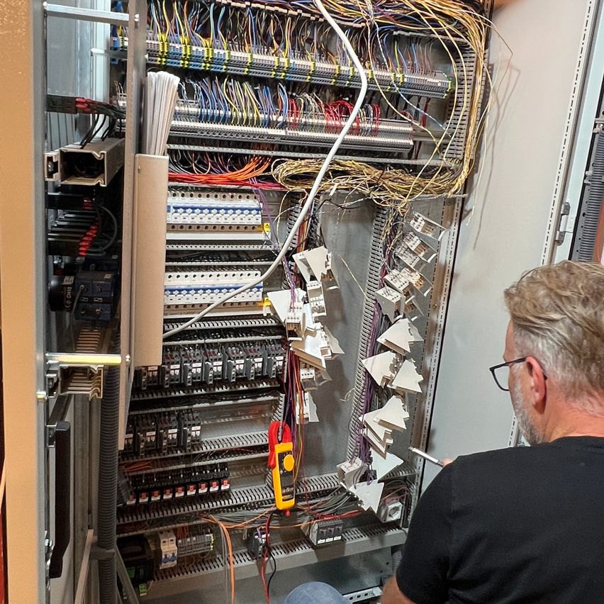 Ein Elektriker arbeitet an einer großen Leiterplatte, umgeben von verschiedenen Kabeln und Komponenten. Er trägt eine Brille und ein schwarzes Shirt.