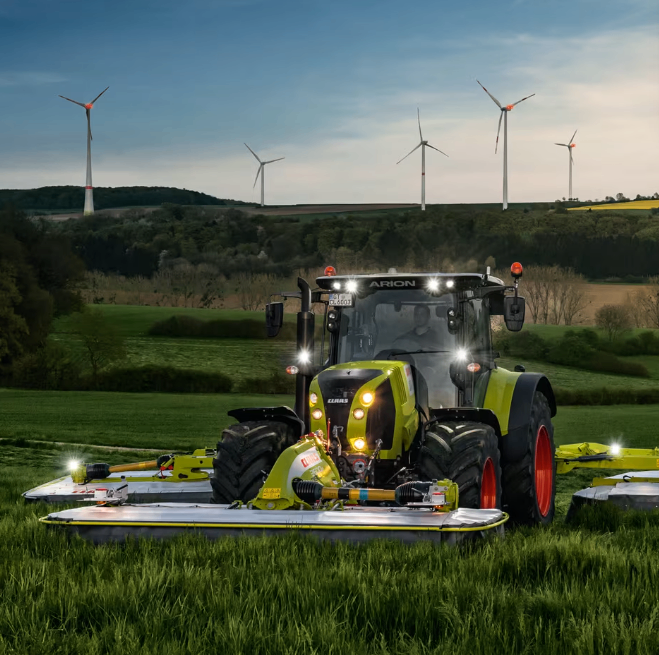 Ein grüner Traktor mit eingeschalteten Lichtern steht auf einem Grasfeld mit Windrädern im Hintergrund.
