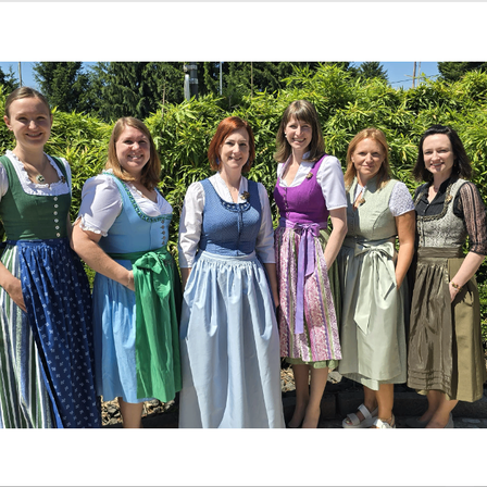 Sechs Frauen stehen zusammen in traditionellen Kleidern und lächeln für ein Foto. Sie tragen farbenfrohe Kleidung mit unterschiedlichen Mustern. Sie stehen vor einem Busch mit Bäumen im Hintergrund.