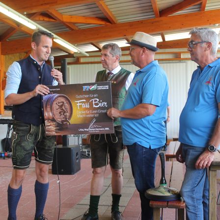 Vier Männer stehen in einem Pavillon. Ein Mann hält ein Mikrofon und überreicht einem anderen Mann einen Gutschein für ein Fass Bier. Die anderen beiden Männer schauen auf den Gutschein.