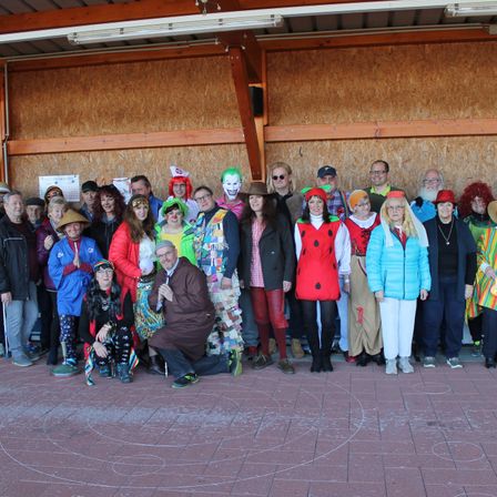 Eine Gruppe von Erwachsenen in bunten Kostümen, einige mit Hüten, posieren für ein Foto in einem Raum mit Ziegelboden.