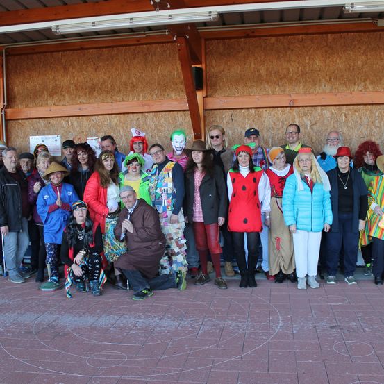 Eine Gruppe von Erwachsenen in bunten Kostümen, einige mit Hüten, posieren für ein Foto in einem Raum mit Ziegelboden.