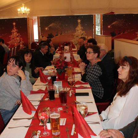 Eine Gruppe von Menschen sitzt an einem Tisch mit Getränken, einem Weihnachtsbaum und einer Weihnachtsdekoration an der Wand.