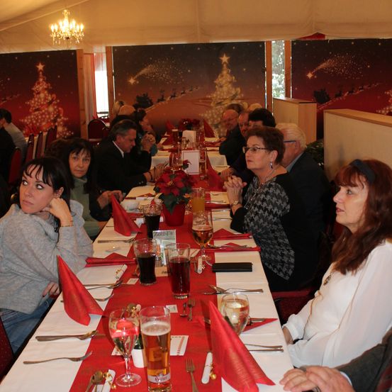 Eine Gruppe von Menschen sitzt an einem Tisch mit Getränken, einem Weihnachtsbaum und einer Weihnachtsdekoration an der Wand.