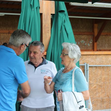 Drei Menschen stehen in einem Freiluftbereich, wahrscheinlich in einem Park, und scheinen zu sprechen. Ein Mann trägt ein weißes Poloshirt mit dem Text "Barood Island" darauf. Die Frau trägt eine Halskette, Ohrringe und einen Armreif und hält eine Flasche in der Hand.