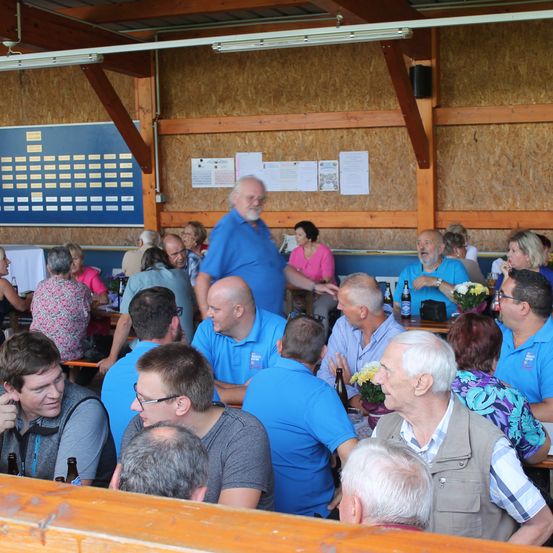 Eine Gruppe von Menschen in einem Saal, einige tragen Brillen und blaue Hemden, einige unterhalten sich, und es gibt Flaschen auf dem Tisch.