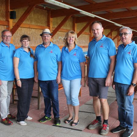 Eine Gruppe von sechs Personen, alle in blauen Hemden mit einem Logo, stehen zusammen in einem Raum mit Holzdecken.