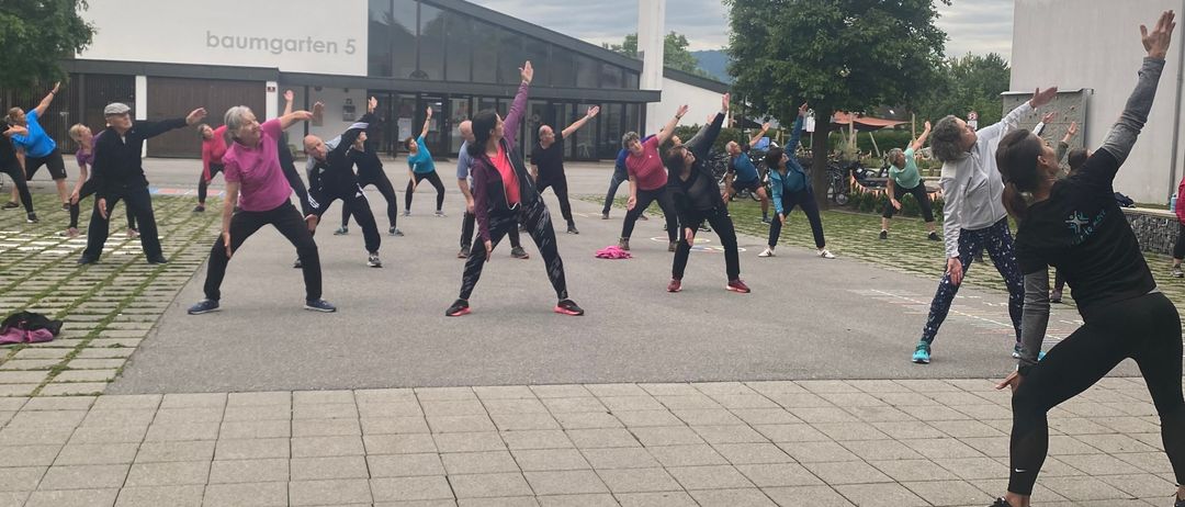 Menschen praktizieren Yoga vor einem Gebäude mit der Aufschrift 'baumgarten 5'. Es ist ein bewölkter Tag mit Bäumen und Pflanzen im Hintergrund.