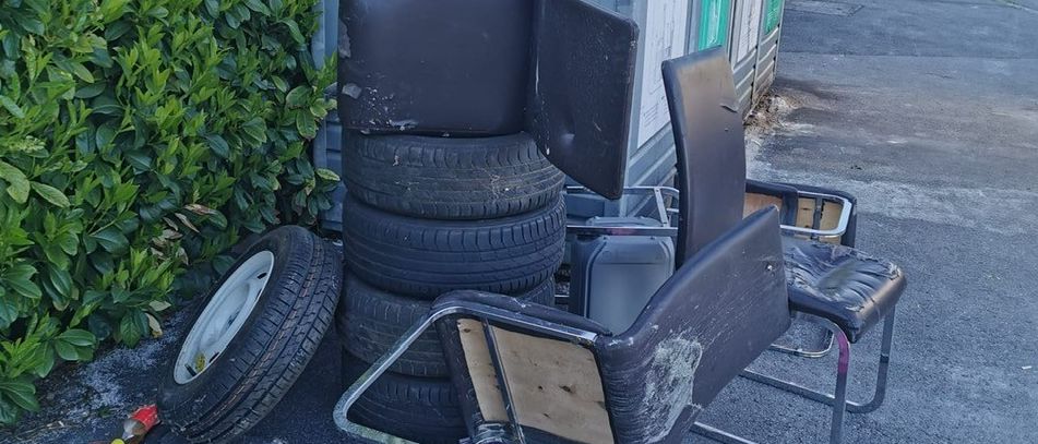 Ein Stapel Reifen und beschädigte Stühle stehen neben einer Recyclingtonne hinter einer Hecke. Eine leere Flasche liegt auf dem Boden.
