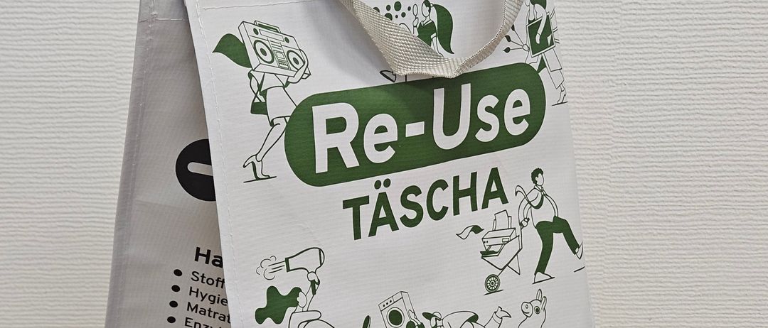 Ein wiederverwendbarer Beutel mit Illustrationen verschiedener Aktivitäten, beschriftet mit 'Re-Use Tasche'. Der Beutel hat Text und Logos, auf einem Holzboden gefaltet.