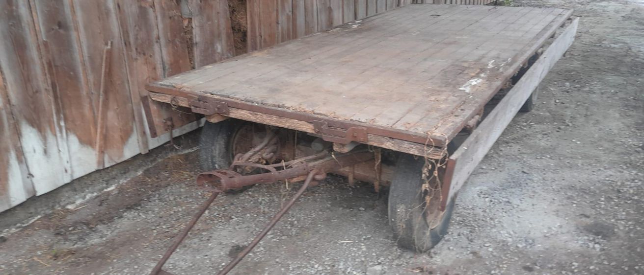 Ein alter hölzerner Anhänger mit rostigen Rädern steht auf Kies neben einer Holzscheune mit einem Loch in der Wand.