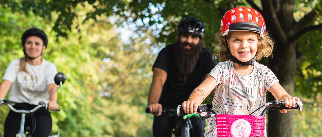 Drei Radfahrer fahren auf einem Kiesweg, umgeben von Bäumen. Ein junges Mädchen mit einem rosa Helm fährt vorne, gefolgt von einem bärtigen Mann und einer Frau.