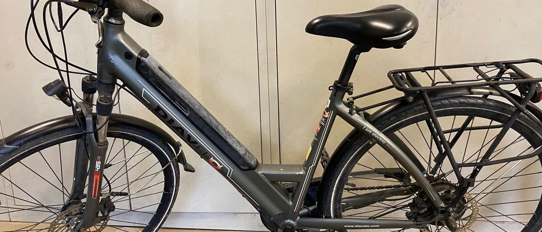 Ein Elektrofahrrad mit schwarzem Sattel und Lenker steht vor einer weißen Wand. Das Fahrrad hat einen schwarzen Gepäckträger hinten und eine vordere Federung.