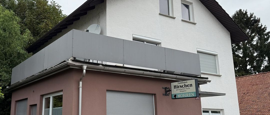 Das Bild zeigt ein zweistöckiges Gebäude mit einem grauen Dach und rosa Wänden. Das Gebäude hat mehrere Fenster und ein kleines Schild mit den Worten Hirschen und Mohren.