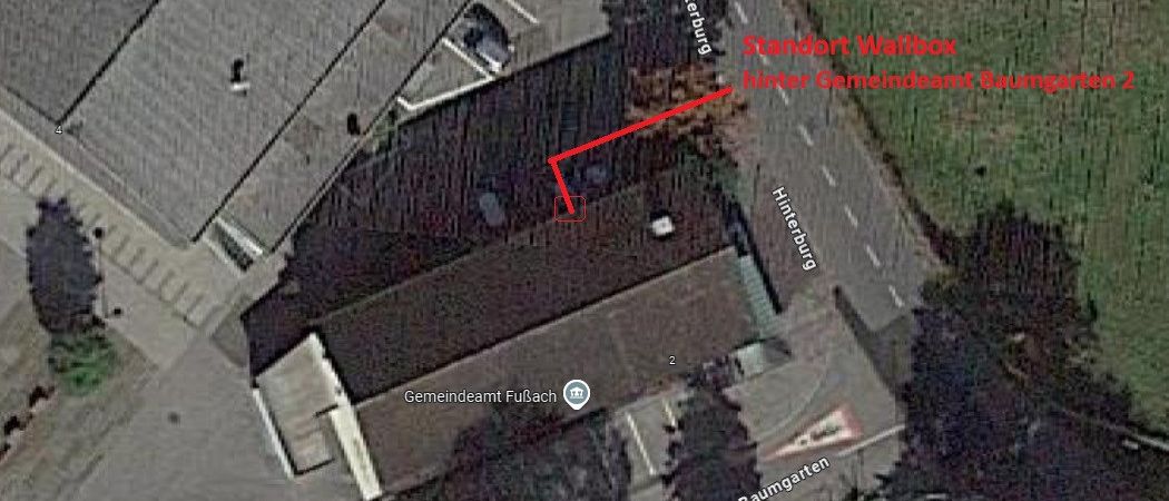 Luftaufnahme eines Gebäudes mit der Aufschrift 'Gemeindeamt Fußach', mit einem roten Pfeil, der auf einen Wallbox-Standort hinter dem Gebäude zeigt. Das Gebiet umfasst Straßen, Parkplätze und Bäume.