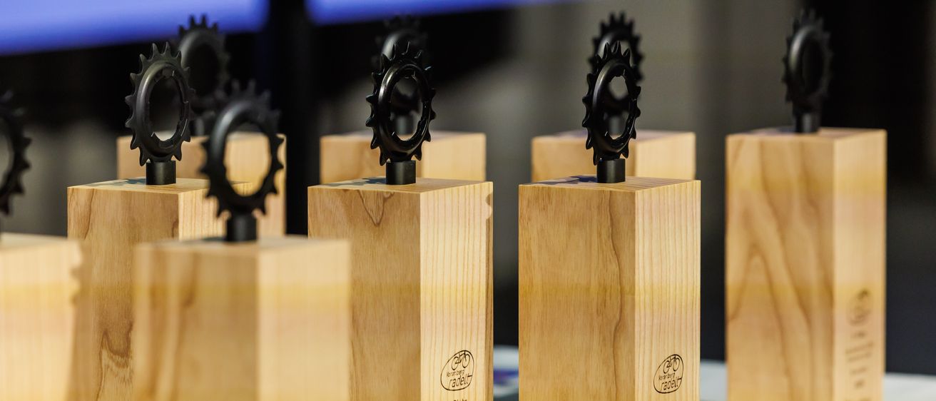 Mehrere hölzerne Trophäen mit schwarzen zahnradähnlichen Oberteilen werden auf einer weißen Oberfläche ausgestellt. Jede Trophäe hat ein schwarzes Zahnrad oben und der hölzerne Sockel ist graviert.