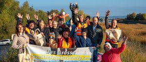 A group of people in safety vests and hats stand with a banner in a grassy area. Some hold bags with the word 'Fussach'. They pose for a photo with a body of water in the background.