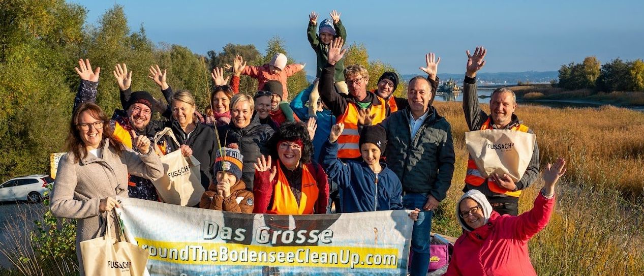 Eine Gruppe von Menschen in Warnwesten und Mützen steht mit einem Banner in einem Grasbereich. Einige halten Taschen mit dem Wort 'Fussach'. Sie posieren für ein Foto mit einem Gewässer im Hintergrund.