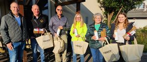 Six people stand outside a house, each holding a bag and a gift box. They are smiling and appear to be posing for a photo.