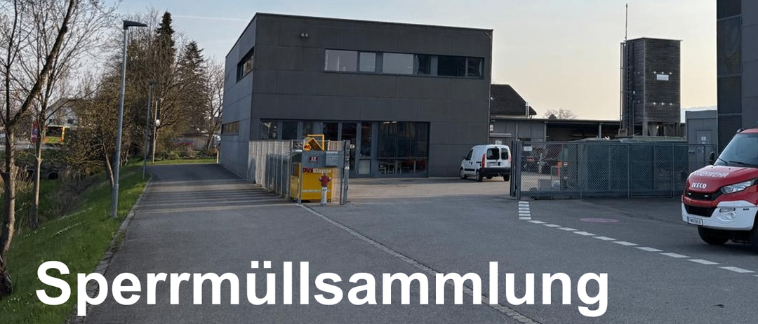 Ein Gebäude mit mehreren Fenstern steht in einem offenen Bereich, mit einem weißen Van vor der Tür. Ein gelber Container steht in der Nähe eines Metallzauns. Bäume und ein Haus sind im Hintergrund sichtbar.