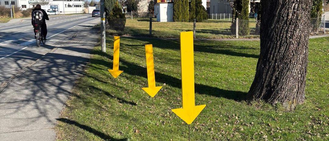 Ein Radfahrer fährt auf einem Bürgersteig mit drei gelben Pfeilen, die nach oben zeigen. Der Bürgersteig grenzt an einen Rasenbereich mit einem großen Baumstamm. Im Hintergrund sind Gebäude und Straßenlaternen zu sehen.