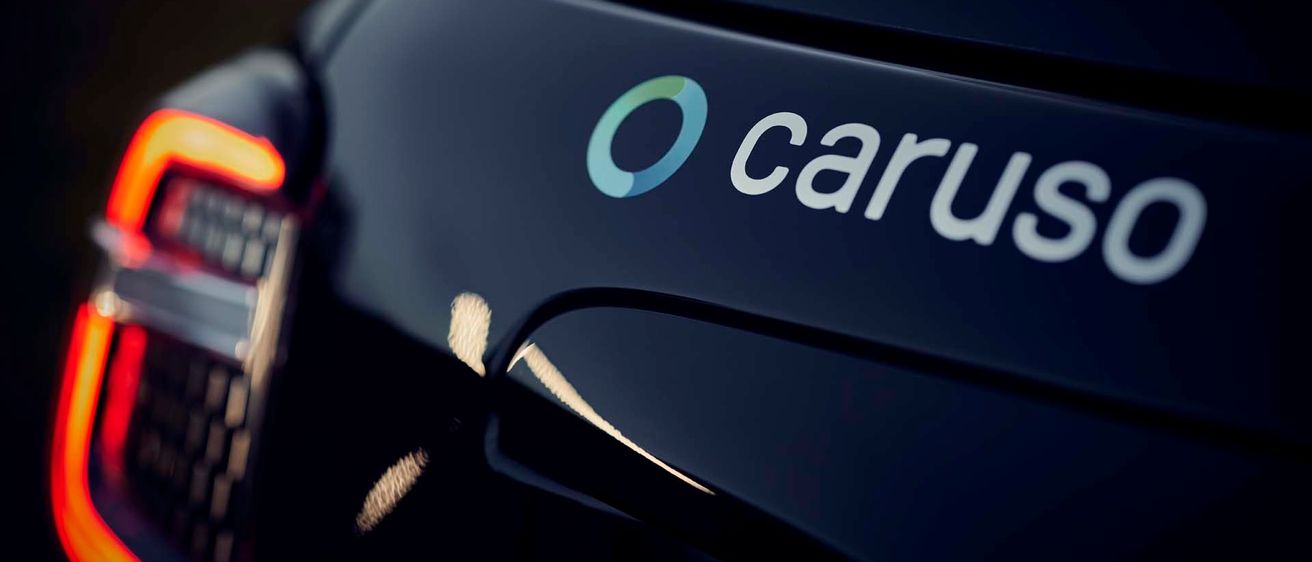 Ein Nahaufnahme des Hecks eines schwarzen Autos mit dem Logo 'O carus' in Weiß auf dunklem Hintergrund. Die Rückleuchte des Autos befindet sich auf der linken Seite.