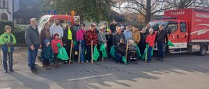 Eine Gruppe von Menschen ist für ein Foto versammelt, in Winterkleidung, einige halten Stöcke und grüne Taschen. Ein Baby in einem Kinderwagen steht in der Mitte. Hinter ihnen stehen zwei Rettungsfahrzeuge.