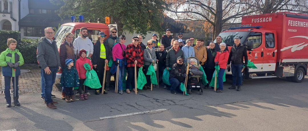 Eine Gruppe von Menschen ist für ein Foto versammelt, in Winterkleidung, einige halten Stöcke und grüne Taschen. Ein Baby in einem Kinderwagen steht in der Mitte. Hinter ihnen stehen zwei Rettungsfahrzeuge.