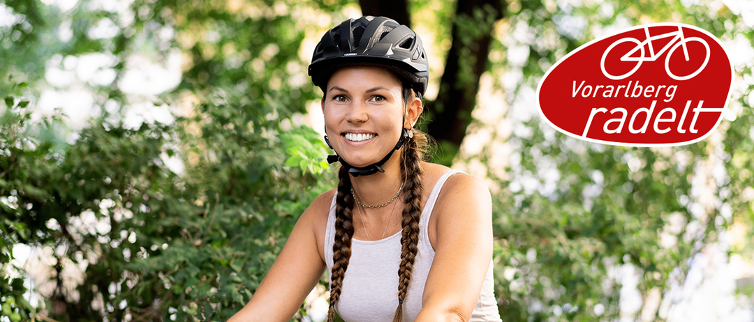 Eine Frau mit geflochtenen Haaren und einem schwarzen Helm fährt Fahrrad im Wald. Sie trägt ein weißes Tanktop und hat ein Tattoo auf dem linken Arm.