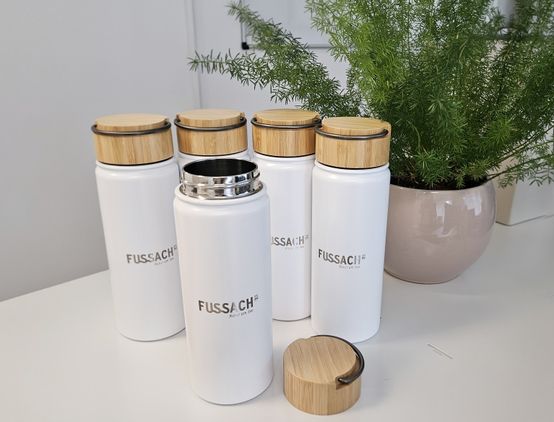 Fünf weiße Thermoflaschen mit Bambusdeckeln sind auf einer weißen Oberfläche ausgestellt. Die Flaschen haben ein schlankes Design und sind mit dem Wort 'Fussach' beschriftet. Ein Topf mit Pflanzen befindet sich dahinter.