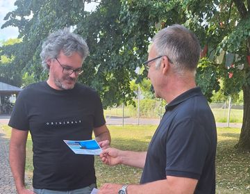 Zwei Männer stehen unter einem Baum. Einer hält einen Flyer und der andere schaut ihn an. Der Mann rechts trägt eine Brille und eine Uhr.