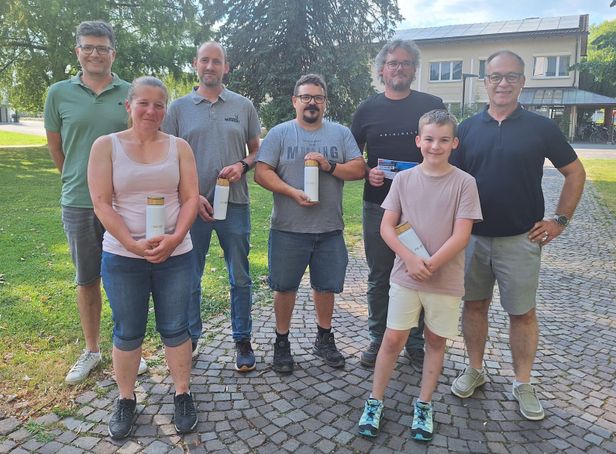 Eine Gruppe von Menschen steht draußen und lächelt, während sie Tassen halten. Dahinter befindet sich ein Gebäude mit Solarmodulen und Bäumen.