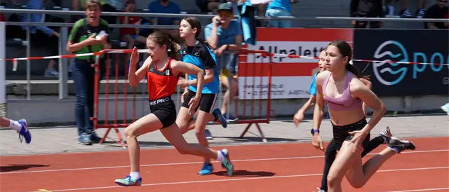 Eine Gruppe von Mädchen läuft auf einer Laufbahn. Zuschauer beobachten vom Rand aus. Die Bahn hat gelbe Linien und ein Sponsorenlogo.