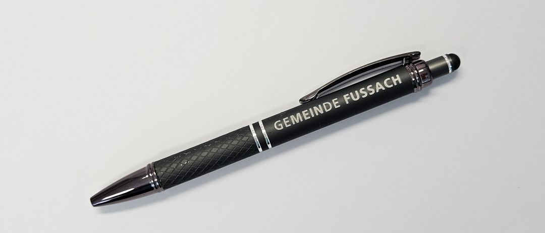 Ein schwarzer Stift mit silbernen Akzenten und dem Wort 'Gemeinde Fussach' darauf geschrieben. Der Stift liegt auf einer weißen Oberfläche.