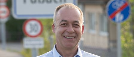 Ein Mann in einem hellblauen Hemd mit einem Lächeln steht auf einer Straße mit einem Geschwindigkeitsbegrenzungsschild dahinter.